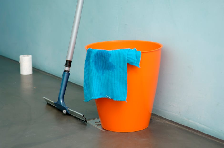 Close-up of an orange plastic bucket with a blue cleaning cloth draped over its side, placed on a smooth, dark grey floor next to a wall painted in a mint green color. A mop with a blue handle and a f