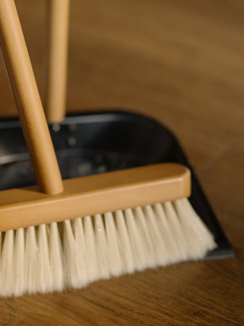 Close-up of an orange plastic bucket with a blue cleaning cloth draped over its side, placed on a smooth, dark grey floor next to a wall painted in a mint green color. A mop with a blue handle and a f