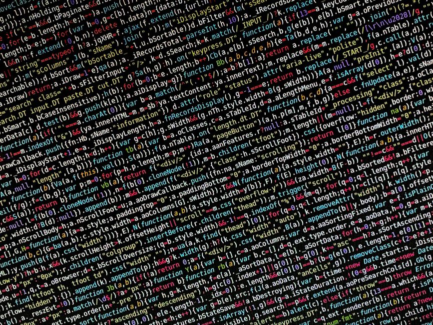 A person seen from behind working on a laptop with a black screen displaying colorful lines of code. The workspace includes a white desk, a closed black monitor, and a black keyboard. The background f